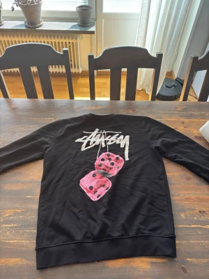Svart Stüssy sweatshirt med tärningar - Cool svart sweatshirt från Stüssy med deras ikoniska logga i vitt på bröstet och en stor print på ryggen med två rosa tärningar. Tröjan har rund halsringning och ribbade muddar vid ärmslut och nederkant. Perfekt för en streetwear-look.