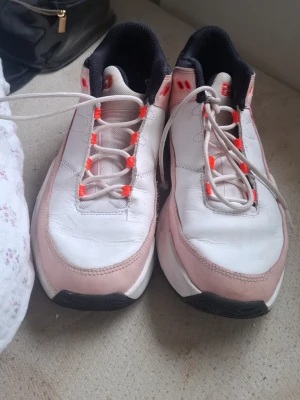 Vita och rosa Air Jordan sneakers - Snygga Air Jordan sneakers i vitt och ljusrosa med svarta detaljer och orange accenter på snörningen. Skorna har en klassisk låg siluett och är tillverkade i läder och textil. Perfekta för dig som vill ha en sportig och trendig look.