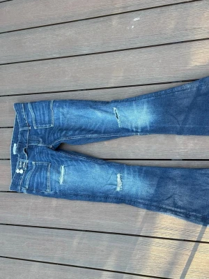 Blå slitna jeans från Gina Tricot - Säljer ett par blå jeans från Gina Tricot med slitningar och ljusa tvättade partier. Jeansen har bootcut-modell, klassiska fickor och dubbla knappar i midjan. Perfekta för en avslappnad och trendig look.