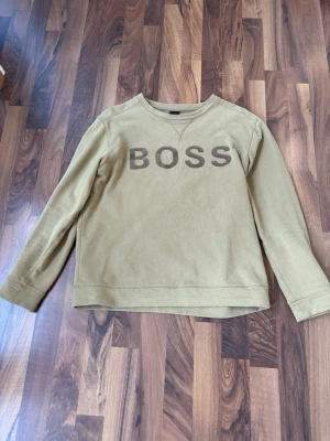 Beige sweatshirt från BOSS - Snygg beige sweatshirt från BOSS med stor logga framtill. Tröjan har rund halsringning, långa ärmar och är gjord i mjukt bomullsmaterial. Perfekt för en avslappnad och trendig stil.