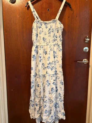 Vit maxiklänning med blå blommor - Lång maxiklänning med smala axelband och smock upptill. Klänningen är vit med blått blommönster och har volangdetaljer nedtill. Tillverkad i ett lätt och luftigt tyg, perfekt för varma dagar. Romantisk och somrig stil.