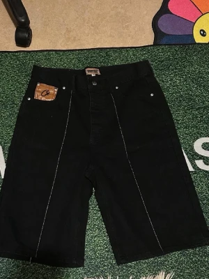 Svarta denimshorts från corteiz - Säljer ett par svarta denimshorts med vita kontrastsömmar och unika stjärndetaljer på bakfickorna. Shortsen har fem fickor, varav en med brun patch framtill, och är tillverkade i ett kraftigt jeanstyg. Perfekta för dig som gillar streetwear och vill sticka ut.