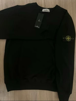 Stone Island tröja svart - Svart sweatshirt från Stone Island med klassisk rund hals och ikonisk logopatch på vänster ärm. Tröjan har ribbade muddar vid ärmslut och nederkant. Perfekt för en clean och stilren look.
