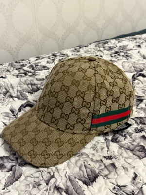 Beige keps med GG-mönster från Gucci - Säljer en beige keps från Gucci med det klassiska GG-monogrammet över hela kepsen. På sidan finns en grön och röd rand som ger extra lyxig känsla. Kepsen har böjd skärm och är tillverkad i ett slitstarkt tygmaterial. Perfekt för dig som vill sticka ut med stil.
