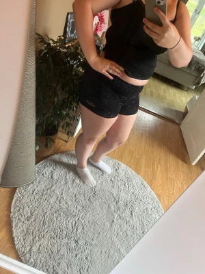 Svarta träningstights shorts - Säljer ett par svarta träningstights shorts med diskret mönster och tight passform. Perfekta för gymmet eller löprundan. De har en hög midja och är gjorda i ett stretchigt syntetmaterial som sitter skönt på kroppen.