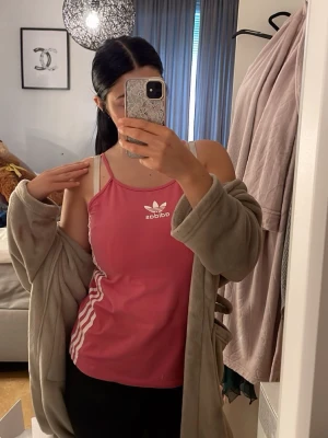 Rosa linnetopp från Adidas - Säljer en rosa linnetopp från Adidas med tunna axelband och vita detaljer. Toppen har Adidas-logga framtill och klassiska vita ränder längs sidorna. Perfekt för en sportig och avslappnad stil.