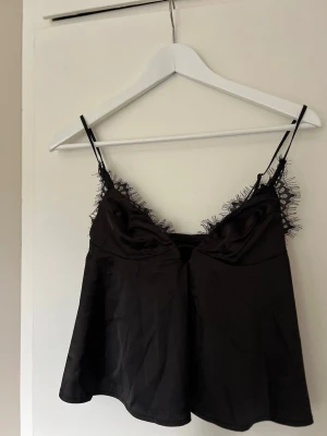 Svart satin body med spetsdetaljer - Snygg svart topp i satin med tunna axelband och spetsdetaljer längs urringningen. Materialet är glansigt och mjukt, vilket ger en lyxig känsla.