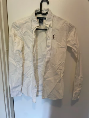 Vit skjorta från Ralph Lauren - Klassisk vit skjorta från Ralph Lauren med broderad logga på bröstet. Skjortan har lång ärm, knappar framtill och en tidlös design som passar till det mesta. Tillverkad i bomull för en skön känsla.