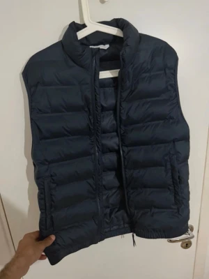 Mörkblå dunväst från ZARA - Snygg mörkblå dunväst från ZARA med hög krage och dragkedja framtill. Västen har två sidofickor och quiltad design som ger en modern look. Perfekt att slänga över en hoodie eller tröja för extra värme. Materialet är lätt och smidigt.