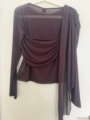 Long sleeve shawl top - Säljer en unik chocolate plum topp med lång ärm och asymmetrisk design. Toppen har snygg drapering över ena axeln och är tillverkad i ett tunt, transparent material som ger en cool och trendig look. Detta skulle passa snyggt med en raka vita byxor!!😍Perfekt för dig som vill sticka ut med din outfit. 💕