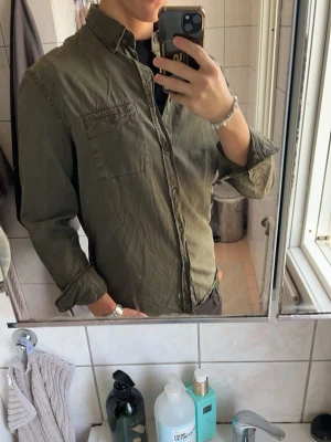 Olivgrön skjorta med bröstficka från Filippa K - Snygg olivgrön skjorta från Filippa K i bomull med klassisk krage och knäppning framtill. Skjortan har en bröstficka på vänster sida samt långa ärmar med manschetter. En stilren och tidlös modell som passar lika bra till vardags som för en mer uppklädd, avslappnad look. Bekväm kvalitet och enkel att matcha med resten av garderoben.