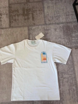 Vit Casa Blanca t-shirt med tryck - Vit t-shirt från Casa Blanca med färgglatt grafiskt tryck i blått, orange och gult både fram och bak. Klassisk rund hals och korta ärmar. Tillverkad i mjuk bomull för en skön känsla.