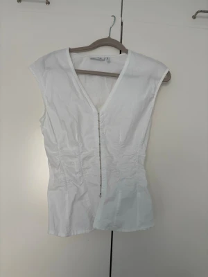 Vit v-ringad blus med dragkedja - Säljer en vit blus med v-ringning och dragkedja framtill. Toppen är ärmlös och har en figurnära passform som framhäver siluetten. Perfekt att styla med jeans eller kjol för en clean look.