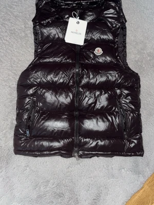 Svart dunväst från Moncler - Snygg svart dunväst från Moncler med quiltad design och glansig finish. Västen har dragkedja framtill, två fickor med dragkedja och Moncler-logga på bröstet. Perfekt för lager-på-lager och streetstyle.