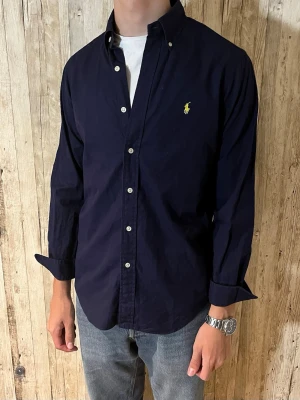 Mörkblå skjorta från Ralph Lauren - Klassisk mörkblå skjorta från Ralph Lauren med knappar framtill och lång ärm. Skjortan har en gul broderad logga på bröstet och button-down krage. Perfekt till jeans eller chinos för en stilren look.
