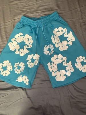 Ljus blåa Denim Tears shorts. - Säljer ett par turkosa shorts med elastisk midja och snörning. Shortsen har flera vita, abstrakta tryck över hela plagget och en bakficka. Perfekta för sommaren och riktigt sköna att bära till vardags. Pris kan diskuteras vid snabb affär. 