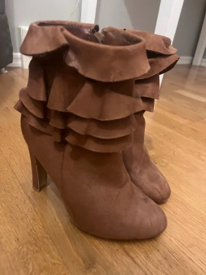 Bruna stövletter med volangdetaljer - Unika bruna stövletter med höga klackar och flera lager volangdetaljer runt skaftet. Tillverkade i ett mockaliknande material och har dragkedja på sidan för enkel på- och avtagning. Perfekta för dig som vill sticka ut med din stil.