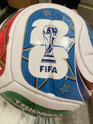 Vit fotboll FIFA Trionda League - Säljer en vit fotboll med färgglada detaljer i blått, grönt och rött. Bollen har FIFA-logga och Trionda League-tryck, samt stjärnmotiv och text på flera språk. Perfekt för matcher eller träning på planen.