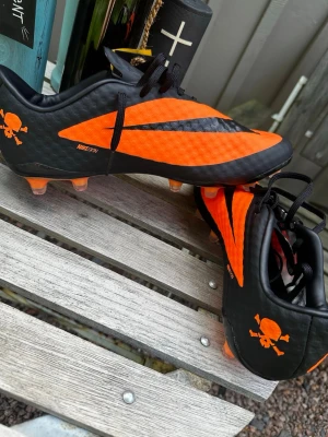 Nike hypervenom phantom regen - Snygga fotbollsskor från Nike i svart och orange med dödskalle-tryck på hälen. Skorna har plastdobbar och en låg profil för bra bollkontroll. Tillverkade i syntetmaterial med cool design och tydliga Nike-loggor. Perfekta för dig som vill sticka ut på planen.