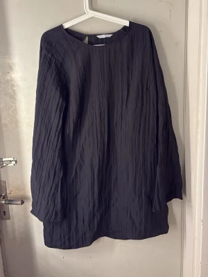 Svart långärmad kortklänning - En svart, långärmad kortklänning i ett krinklat tyg med rund halsringning. Klänningen har en enkel och stilren design utan synliga detaljer eller mönster, vilket gör den lätt att styla till olika looks.
