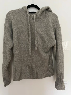 Grå stickad hoodie med huva - Mysig grå stickad hoodie.  Tröjan har ribbstickade muddar vid ärmslut och nederkant, samt en avslappnad passform. Perfekt för en chill och stilren look.