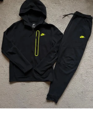 Svart Nike tech suit  - Säljer ett svart Nike träningsset med zip-hoodie och matchande byxor. Setet har neongula Nike-loggor och en snygg detalj med gul dragkedja på bröstet. Perfekt för gymmet eller chill. Mjukt material och skön passform.                                          Storlek S på hoodien och storlek M på byxor!!