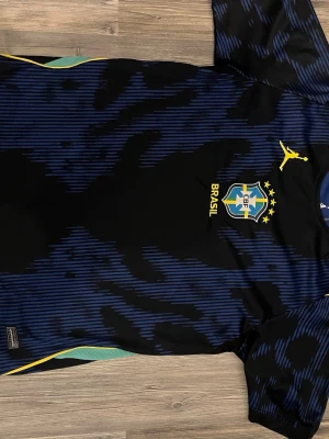 Blå Brasilien fotbollströja - Säljer en blå och svart fotbollströja med Brasilien-emblem och gula detaljer. Tröjan har ett randigt mönster och gula inslag vid axlar och sidor. Perfekt för dig som älskar fotboll och vill stötta Brasilien på match eller träning.