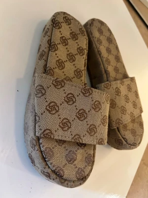 Beige tofflor med monogrammönster - Säljer ett par beige tofflor med bred rem och brunt monogrammönster över hela skon. Tofflorna har öppen tå och är tillverkade i ett tygmaterial som ger en avslappnad och trendig look. Perfekta för att chilla hemma eller på semestern.