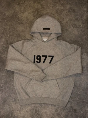 Hoodie Fear of God ESSENTIALS 1977 Grey 👕 - This Fear of God ESSENTIALS 1977 hoodie in heather grey features a prominent "1977" print on the chest and a signature rubberized Essentials patch on the hood. Crafted from a comfortable cotton blend, it includes a classic kangaroo pocket and raglan sleeves.  ⭐️ Condition : 9/10 🎨 Color : Heather Grey 📦 Fast and careful shipping.