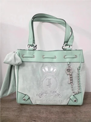 Mintgrön handväska Juicy Couture daydreamer tote  - Snygg mintgrön handväska från Juicy Couture i mocka och skinnimitation. Väskan har dubbla handtag, silverfärgade detaljer, en dekorativ nyckelberlock och ett broderat emblem framtill. Stor rosett på sidan ger extra attityd.