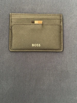 Svart kortfodral från BOSS - Snyggt svart kortfodral från BOSS i stilren design. Tillverkat i strukturerat läder med flera kortfack både fram och bak. Diskret BOSS-logga framtill och en liten detalj i beige, svart och orange upptill. Perfekt för dig som vill ha koll på korten utan att ta plats.