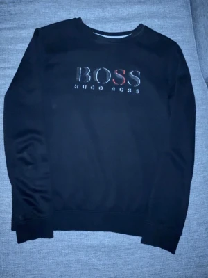 Svart sweatshirt från Hugo Boss - Svart sweatshirt från Hugo Boss med stor BOSS-logga i vitt och rött framtill. Klassisk rund halsringning och långa ärmar. Tröjan har ribbade muddar vid ärmslut och nederkant. Perfekt för en stilren och avslappnad look.