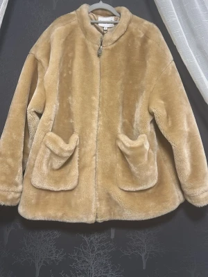 Beige fuskpälsjacka från NA-KD - Snygg beige fuskpälsjacka från NA-KD med dragkedja framtill och två stora fickor. Jackan har en rundad krage och är tillverkad i mjukt, fluffigt material som ger en lyxig känsla. Perfekt för att hålla värmen under kalla dagar.