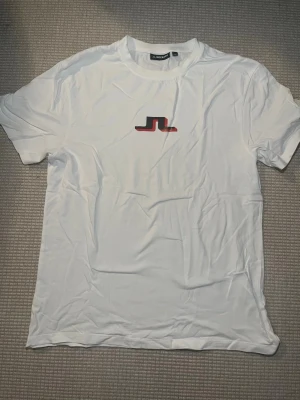 Vit t-shirt från J.Lindeberg - Snygg vit t-shirt från J.Lindeberg med det klassiska JL-logotypen i svart och rött på bröstet. T-shirten har rund halsringning och korta ärmar. Tillverkad i mjukt bomullsmaterial som känns skönt mot huden.