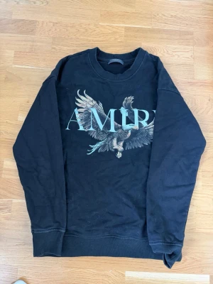 Svart sweatshirt från Amiri med örn - Cool svart sweatshirt från Amiri med stort tryck av en flygande örn och AMIRI-logga på bröstet. Tröjan har rund hals, ribbade muddar och är tillverkad i mjukt bomullsmaterial. Perfekt för dig som gillar streetwear och statement-plagg.