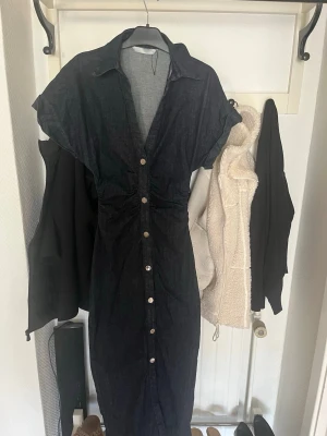 Svart jeansklänning med knappar - Snygg svart jeansklänning med v-ringning och knappar hela vägen framtill. Klänningen har korta ärmar och markerad midja för en smickrande siluett. Perfekt till sommaren och enkel att styla med accessoarer.