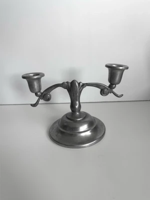 Metallindustri Ystad - Antik tennljusstake med två armar - Stilig tenn-ljusstake från anrika svenska metall- och bruksföremålstillverkaren Metallindustri Ystad. Stilren design med rund bas och två armar med diskreta växt-inspirerade detaljer. Ljusstaken är 19 cm bred och 13 cm hög. Ett stycke svensk designhistoria i ljusstake-form. Nr 47 i serien. Fint skick!