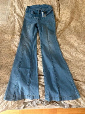 Blå utsvängda jeans med bälte - Säljer ett par blå jeans med hög midja och breda utsvängda ben. Jeansen har ett matchande bälte med stor silverfärgad spänne och markerade sömmar längs benen. Perfekta för dig som gillar retrostil och vill sticka ut.