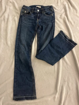 Mörkblå bootcut jeans med kontrastsömmar från Gina teickt - Snygga mörkblå bootcut jeans med kontrastsömmar och rå kant nertill. Jeansen har fem fickor, dekorativa knappar på bakfickorna. Från Gina tricot i storlek och 158. Jeansen är uppsträttade nertill.