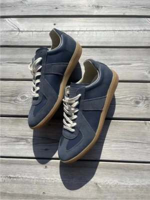 Maison Margiella  - Snygga mörkblå sneakers med vita snören och klassisk gummisula. Skorna har en stilren design med paneler i mocka och textil, perfekta för en avslappnad streetwear-look. Passar till det mesta och är riktigt bekväma att bära.