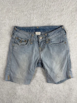 True Religion Becky Super T Jeansshorts W28 Ljus denim - Säljer ett par ljusblå jeansshorts med rå, avklippt kant och klassiska fem fickor. Shortsen har knappgylf och slitna detaljer för en avslappnad look. Perfekta för varma dagar och enkel att matcha med allt.