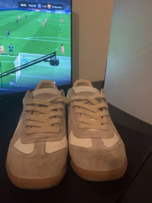 Vita och beige maison margiela skor - Snygga sneakers i vitt och beige med ovandel i läder och mocka. Skorna har klassisk låg profil och gummisula i brunt. Perfekta för en clean och stilren look. Matcha till jeans eller shorts för en avslappnad vibe.