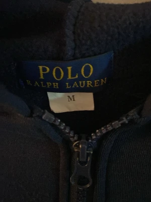 Mörkblå hoodie från Polo Ralph Lauren - Säljer en mörkblå hoodie från Polo Ralph Lauren i storlek M. Hoodien har dragkedja framtill, huva och ribbade muddar vid ärmslut och nederkant. Tillverkad i mjukt material som känns skönt mot huden. Perfekt för en avslappnad och stilren look.