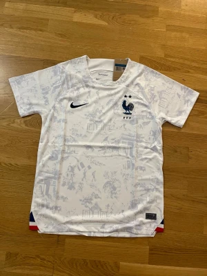  Frankrike fotbollströja - Säljer en vit fotbollströja från Nike med Frankrikes landslagsmärke och två stjärnor på bröstet. Tröjan har diskreta ljusblå mönster, korta ärmar och rund hals. Perfekt för dig som vill stötta Les Bleus på match eller träning.            Fraktar samma eler dagen efter du köpt!!! Endast en kvar i lager!!!