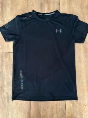 Svart träningströja från Under Armour – Perfekt för gymmet - Säljer en snygg och teknisk träningströja från Under Armour. Det är en stilren svart tröja med snygga, reflekterande detaljer (iridescent effekt) på logotypen och trycket, vilket ger en schysst look både på gymmet och till vardags.  Märke: Under Armour  Modell: Träningströja/Performance shirt  Färg: Svart med reflekterande detaljer 