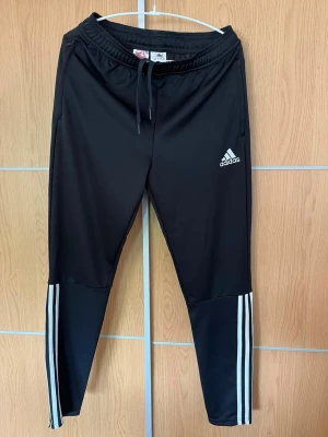 Svarta träningsbyxor från Adidas - Säljer ett par svarta träningsbyxor från Adidas med vita ränder längs benen och logga på låret. Byxorna har dragsko i midjan och är tillverkade i ett lätt, stretchigt material som passar perfekt till sport och träning.