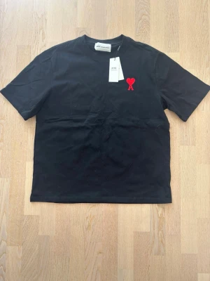 Svart t-shirt från Ami Paris - Svart AA t-shirt från Ami Paris med röd broderad logga i form av ett hjärta och bokstaven A på bröstet. Klassisk rund hals och korta ärmar. Tillverkad i mjuk bomull som känns skön mot huden. Perfekt för en clean och stilren look.                Har använts en gång, i princip helt ny   