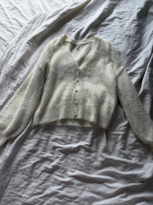 Vit fluffig kofta med pärlknappar - Mysig vit kofta med fluffig känsla och v-ringning. Koftan har långa ärmar och stängs med pärlknappar framtill. Kortare modell som passar perfekt till höga jeans eller kjol. Materialet ser ut att vara en mjuk blandning med mohair eller liknande.