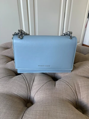 Ljusblå axelremsväska Charles & Keith - Snygg ljusblå handväska från Charles & Keith i slätt konstläder. Väskan har silverfärgad kedjerem och magnetlås. Insidan har ett fack och pryds av ett diskret 'ck'-emblem. Perfekt för dig som gillar stilrena accessoarer.