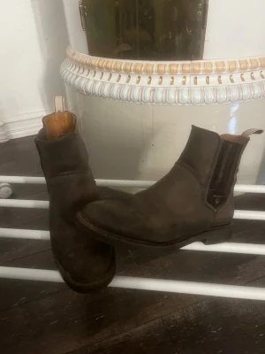 Bruna Chelsea boots från Gant - Snygga bruna Chelsea boots från Gant i mocka med klassisk resår på sidorna och diskret logga i guld. Skorna har en stilren siluett och låg klack, perfekta för en trendig och avslappnad look.